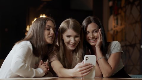 Smiling company of young women in a cafe communicate using a smartphone with a friend. The ladies are discussing among themselves. Friendship concept
