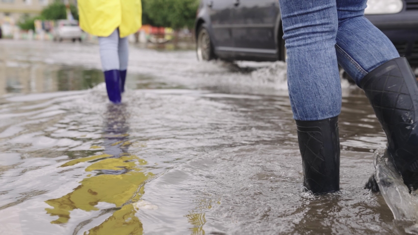 Girls in raincoats and rubber boots walk along road flooded with torrential rains, their feet walk through puddles city, splashing water to the sides, the flood is on street, car is driving on water | Shutterstock HD Video #1082117882