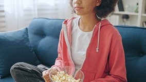 Relaxed teen girl eating popcorn on the sofa, watching comedy at home, weekend