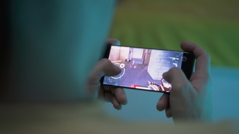 From above anonymous male gamer using cellphone to play shooter at night at home
