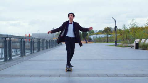 Man in a business suit is rolling on skateboard along embankment, pushing off with his foot, enjoying speed. Guy practicing skateboarding on esplanade. Active, sportive lifestyle, hobby
