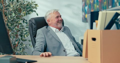Long-time experienced boss of company in old age with gray hair retires promoted on desk there is box with packed things man is happy about rest new position cheers with hands raised in air slaps top