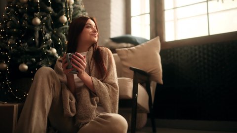 Attractive young woman in the house holding and drinking a cup of tea near the Christmas tree. Decorated Christmas tree.