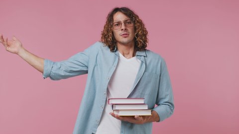 Confused pensive young curly man 20s wears blue shirt white t-shirt wearing translucent pink glasses hold a pile of books spreading hand isolated on pastel plain light pink background studio portrait