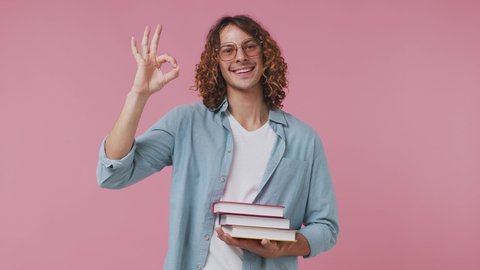 Smiling young curly man 20s wears blue shirt white t-shirt showing okay ok zero finger gesture wearing glasses holding a pile of books isolated on pastel plain light pink background studio portrait