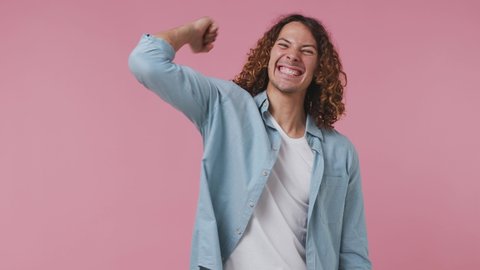 Excited jubilant overjoyed fun young curly man 20s wears blue shirt white t-shirt doing winner gesture celebrate clenching fists say yes isolated on pastel plain light pink background studio portrait
