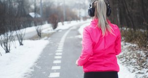 Running woman, headphone girl runner on the snow in winter sunny day. Female in pink sportswear jacket fitness training outdoors. Sport concept, leisure and freedom. 4K slow motion video