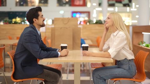 Side view of beautiful young couple drinking coffee and talking sitting at table in mall after shopping in store. Stylish bearded man and blonde woman relaxing after shopping in cafe, slow motion.