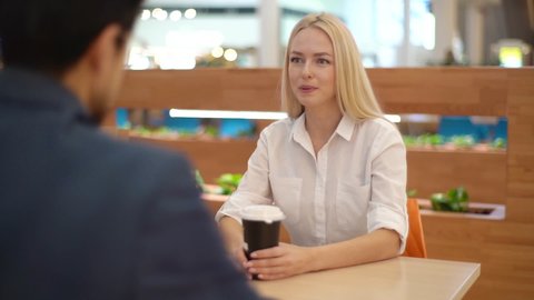 Close-up view from back to cheerful blonde young woman talking with unrecognizable man sitting at table in coffee shop. Happy beautiful couple drinking coffee and smiling in cafe at shopping mall.