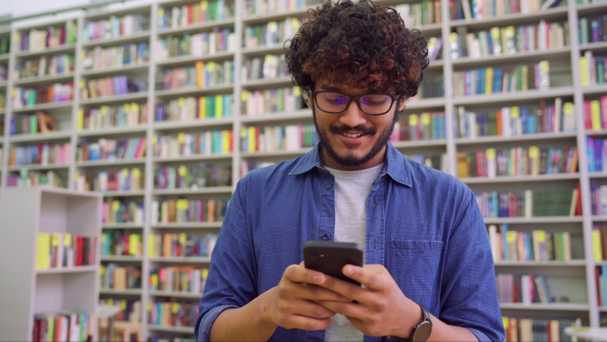 Side view portrait of happy young Indian student chatting by mobile phone in university library. Teen guy wearing denim shirt and eyeglasses using social media application browsing on smartphone Royalty-Free Stock Footage #1082127737