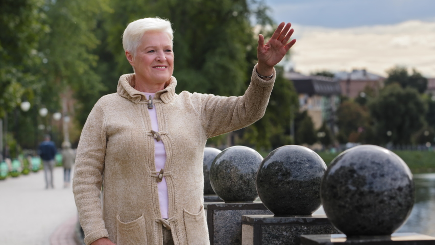 Portrait of smiling aged beautiful female looking away waving hello or goodbye relaxing outdoors in cool weather, happy senior gray-haired woman elderly positive lady in warm clothes carefree concept