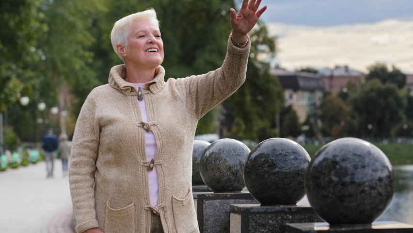Portrait of smiling aged beautiful female looking away waving hello or goodbye relaxing outdoors in cool weather, happy senior gray-haired woman elderly positive lady in warm clothes carefree concept