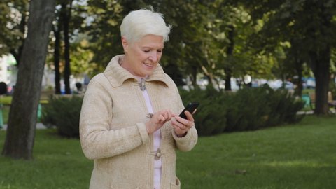 Gray-haired retired woman walking in park in cool weather outdoor, using phone browsing smartphone apps, looking at screen, senior female chatting online, shopping, reading news with mobile device