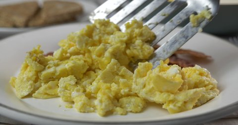 Spooning scrambled eggs onto a plate with bacon and toast