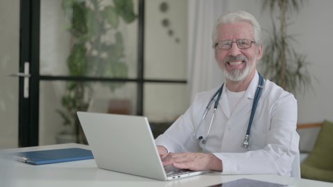 Senior Old Doctor Smiling at Camera while using Laptop
