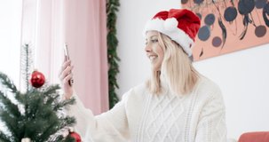 Young smiling woman wearing a santa hat making video call on cosy couch and opens christmas gift in front of selfi cam shot in 4k