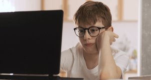 Boy in glasses in the process of online learning with a laptop at home. Schoolboy doing homework. Online training.