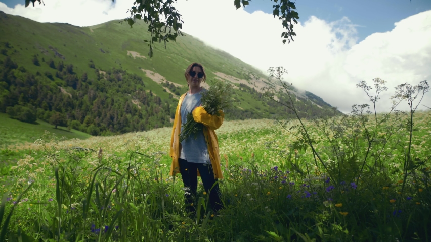 A young woman in a yellow raincoat circles around herself in a meadow with a bouquet of flowers and enjoys the scenery in mountainous areas. Tourism and travel