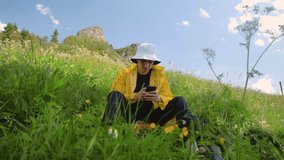 A young man in a yellow raincoat is sitting on a slope and texting on the phone in a mountainous area. Tourism and travel