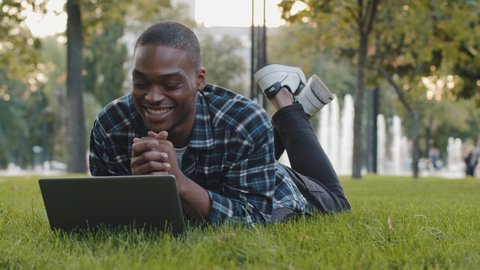 Male student african american guy business man freelancer lies on green grass lawn in park with laptop making video call online conference chat using computer app distant conversation waving hello