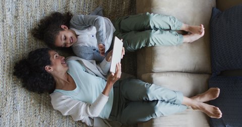 Vertical overhead shot of biracial mother and daughter lying on the floor with their legs on a couch, using a tablet. Family and domestic life concept.