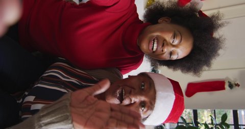 Vertical shot of African American mother and daughter having a video call, wearing Santa Claus hats, waving at the camera. Family and Christmas concept.