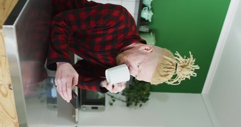Vertical shot of an Albino man leaning over a kitchen isle, drinking coffee and using his smartphone. Domestic life concept.