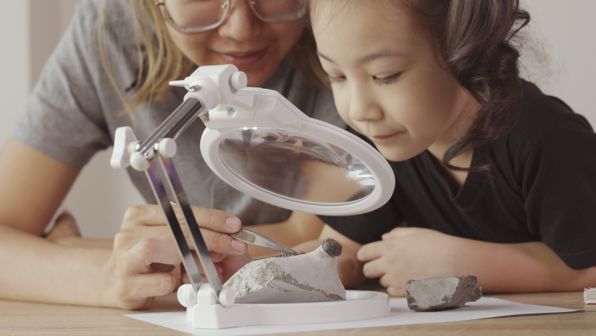 Asian mother and her daughter are learning science experiment at home by observing fossil rock through magnifying glass. It shows concept of family learning with fun, happiness and curiosity.
