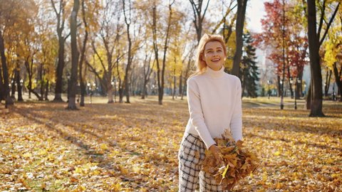 Cute happy young woman swirling with autumn leaves, throwing them in air and dancing in autumn park, slow motion