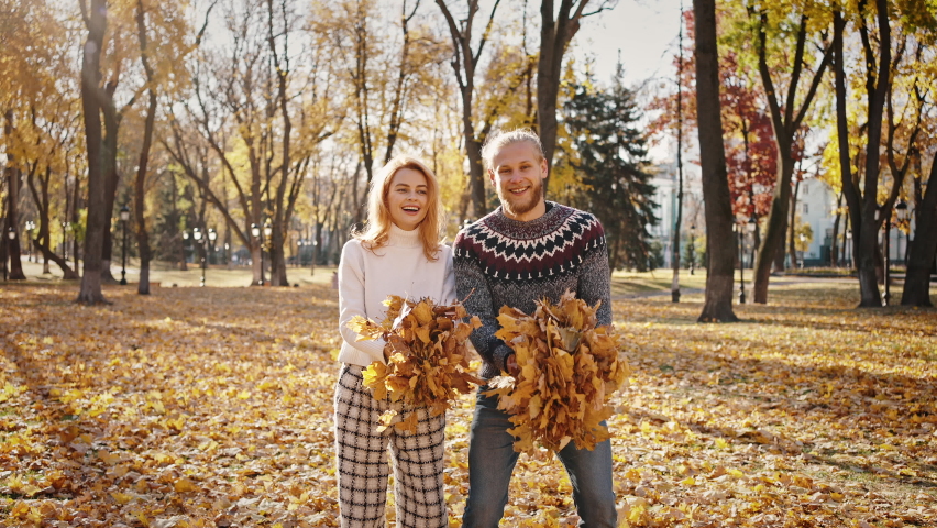 Autumn fun. Young happy man and woman throwing bunches of fallen leaves in park and laughing under rain, fooling around together, slow motion