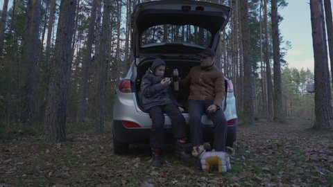 Family picnic during roadtrip in autumn season. Father and son sitting inside car trunk near rural road in autumn forest and drinking tea from thermos.