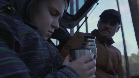 Dad and teen son together sitting on open car trunk and drinking hot tea from steel thermos cup. Concept hiking and family connection.