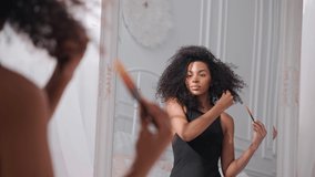 Black woman combing her afro hair, sitting on a bed and looking in mirror. Taking hair care of afro hair in bright white bedroom. High quality 4k footage - Powered by Shutterstock - Get 15% off with code: PIKWIZARD15