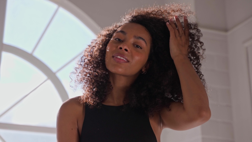 Black woman spraying hairspray, smiling and looking at camera. Taking hair care of afro sitting in bright white bathroom with round window. High quality 4k footage Royalty-Free Stock Footage #1082173040