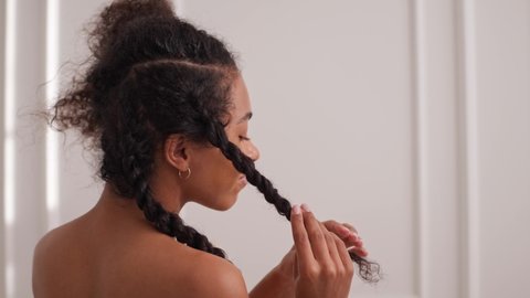 Black young woman twists her hair after bath in bright white room and looks at the camera. Taking care of hair after bath and smiling at the camera. High quality 4k footage