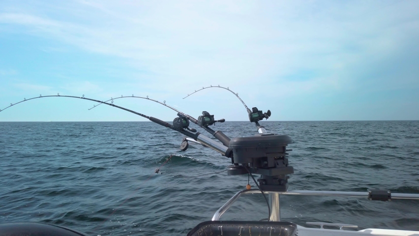 Three fishing rods hooked up with spinners, rigged for fishing, with lines out in the water. The rods are lined up in a row on a small fishing boat. Gentle waves makes the boat rock. Royalty-Free Stock Footage #1082173964