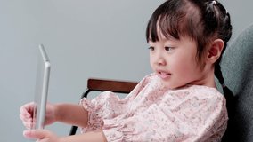 Little asian girl sitting on chair watching online video on tablet, playing video games.