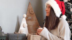 Young beautiful brunette in a Christmas Santa hat, near the eco-tree, looks at Christmas gifts. Family tradition. Gift exchange.