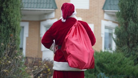 Back view Father Christmas with red bag walking to house scratching head thinking. Santa Clause in red costume with gifts outdoors on backyard. New Year surprise and celebration concept