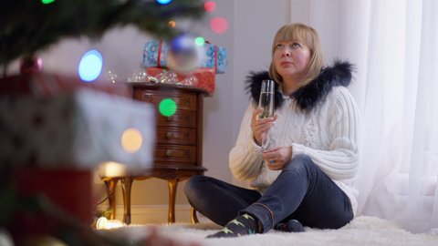 Wide shot portrait sad Caucasian adult woman looking at decorated New Year tree drinking champagne alone. Lonely frustrated depressed lady in living room at home on Christmas eve with alcohol