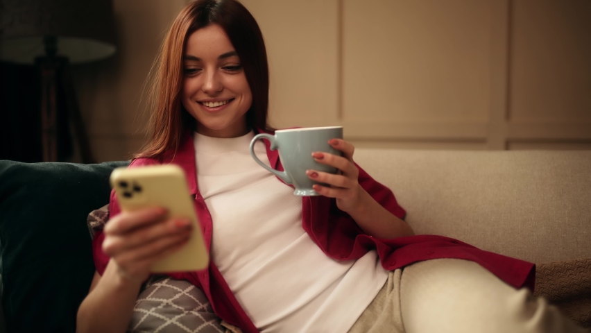 Attractive young woman with cup of tea or coffee using smartphone while sitting on couch. Beautiful girl relaxing while chatting on mobile phone.