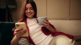 Attractive young woman with cup of tea or coffee using smartphone while sitting on couch. Beautiful girl relaxing while chatting on mobile phone. - Powered by Shutterstock - Get 15% off with code: PIKWIZARD15