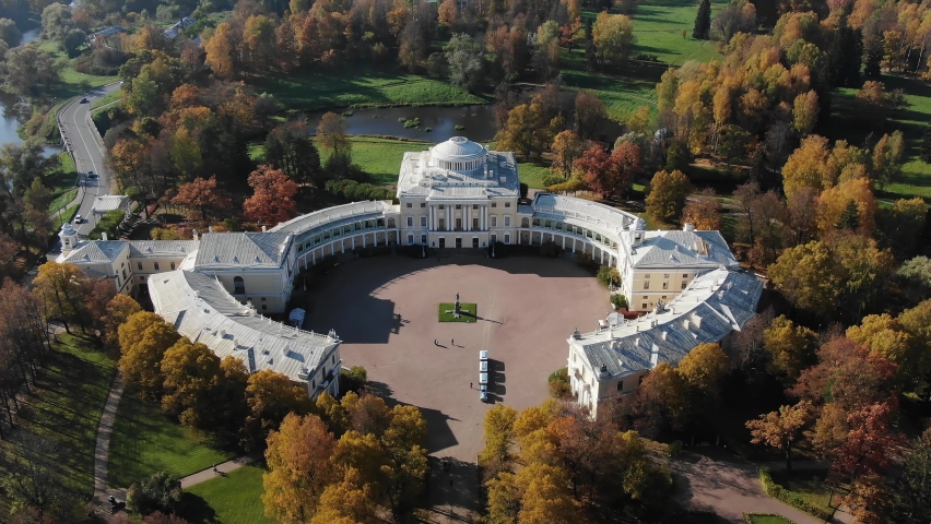 White Pavlovsk historical palace museum among large city park near river with terracotta leaves on trees on sunny autumn day aerial panorama