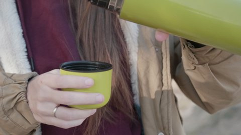 A traveler girl pours tea from a thermos cup during a hike. the girl spills a hot drink into a mug from a green thermos.