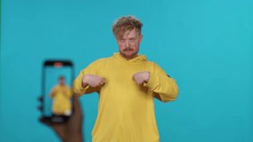 Young guy in yellow hoodie dances on a blue light background while shooting a video on smartphone for social networks, slow motion.