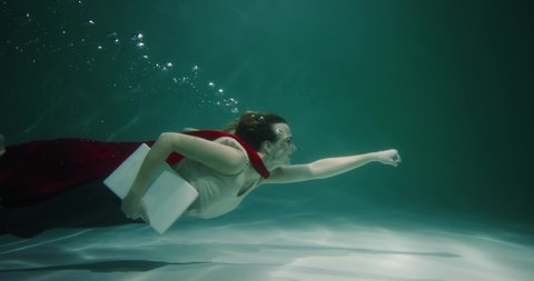 Rushing to help. Beautiful young female client service worker with red superhero cape suit swims underwater slow motion.