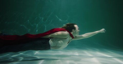 GIRL POWER Beautiful young business woman with red superhero cape swims underwater smiling, rushing to help slow motion.