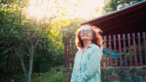 Handheld cinematic shot. Portrait of a mature adult woman in glasses standing against the background of her country house on a windy sunny day.