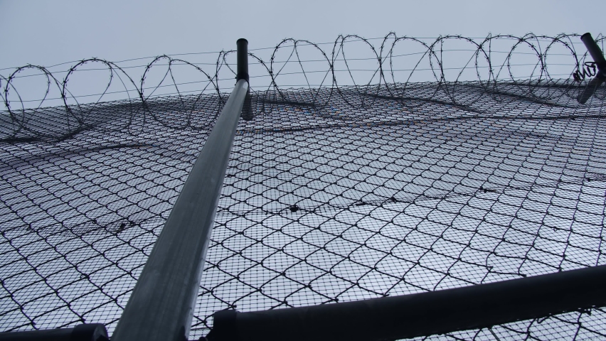 A chain-link fence with barbed wire of the prison or concentration camp with a grey cloudy sky backdrop Royalty-Free Stock Footage #1082208311