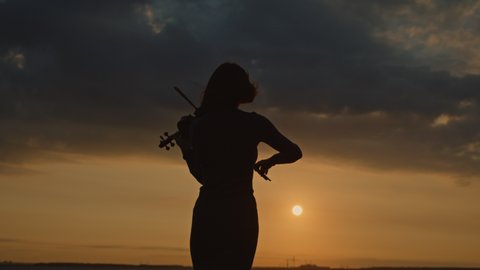 Silhouette of woman from the back with short haircut playing violin outside, in counter sunrise light, getting inspiration of beautiful nature, Slow motion. 